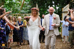 Guests toss confetti over a bride and groom walking a wooded path. The photo captures a candid moment during a woodland wedding celebration.